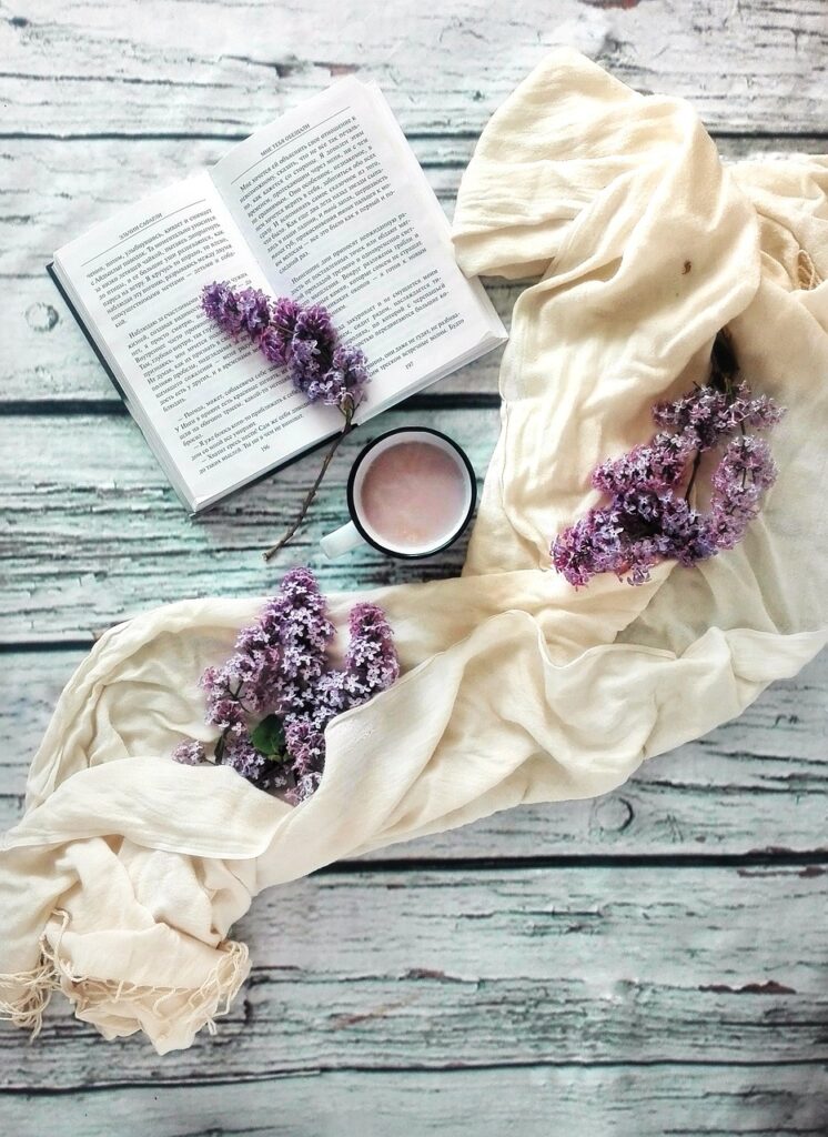 Livre ouvert sur une table en bois, entouré d'un tissu blanc cassé et de branches de lilas. Une tasse de chocolat chaud est disposée au centre.